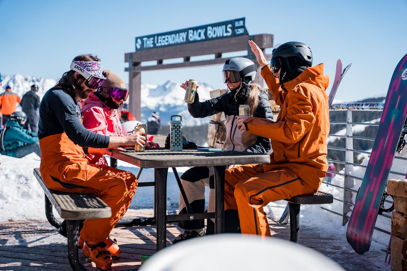 Group of snowboarders sitting at a table with drinks, surrounded by snow and ski equipment.