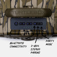 Close-up of a TURTLEBOX electronic device showing the front control panel with labeled callouts for Bluetooth connectivity, 2-BOX stereo pairing, and party mode, over a camouflage-patterned surface.