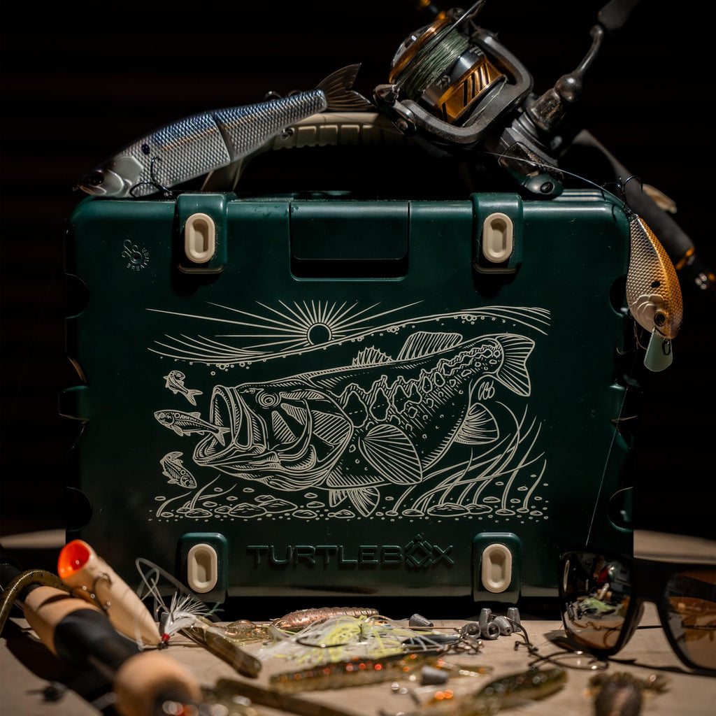 A lifestyle photo of the Bass Artist Series speaker with fish design and fishing gear on a dark background #base-color_bass-artist-series
