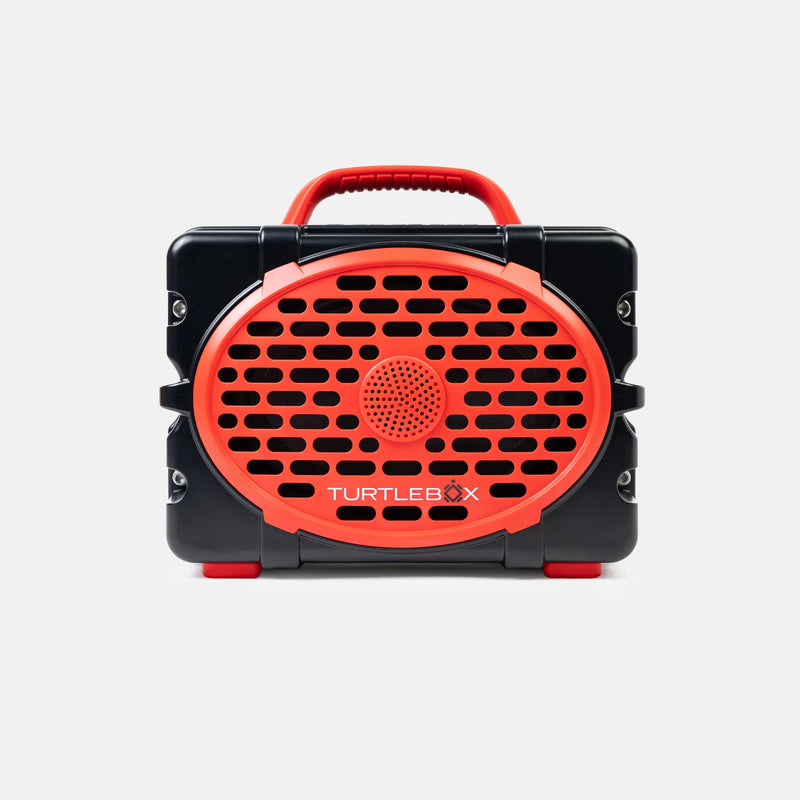 A portable Turtlebox speaker with a black rugged plastic body and bright red accents, including a handle, front grille, and small feet, shown against a light gray background.