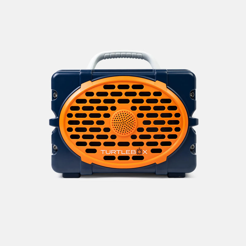 A Turtlebox portable speaker in navy blue and bright orange, featuring a front-facing oval speaker grille with the brand name printed in white, a top carrying handle, and side lugs, shown against a light gray background.