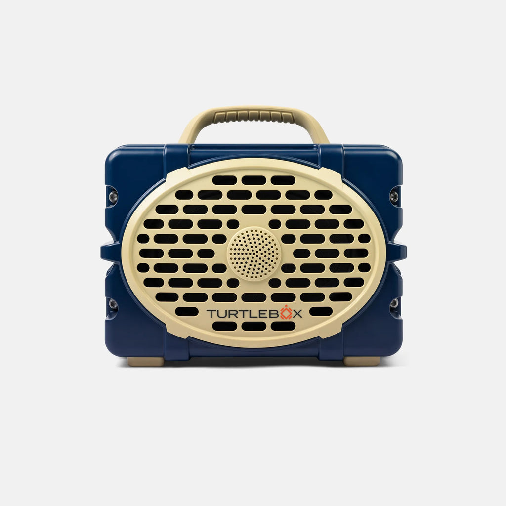 A retro-style portable speaker by TurtleBox, featuring a navy blue body with a cream-colored front grille, handle, and logo, shown against a light gray background.
