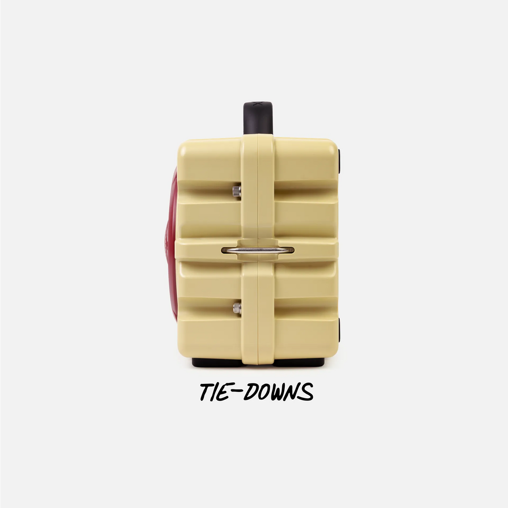 A cream-colored hard-sided suitcase with a vertical strap closure and a red side panel, shown from the back on a light gray background with the text 'TIE-DOWNS' underneath.