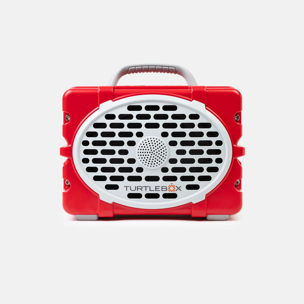 A red portable speaker by TurtleBox with a white oval front grille, black speaker holes, a gray carrying handle, and small gray feet, shown on a light gray background.
