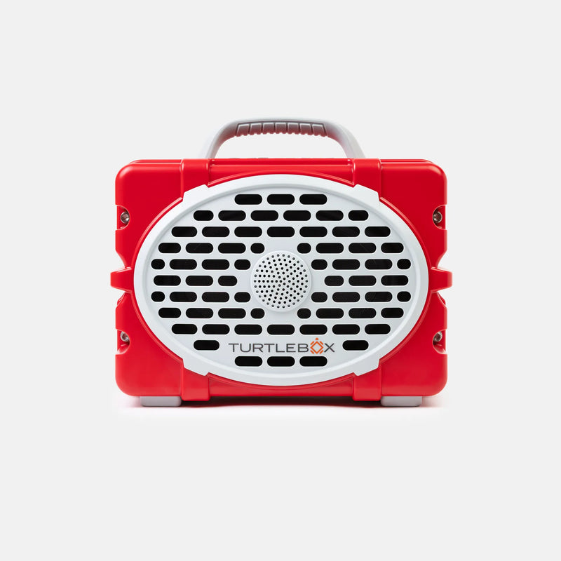 A red portable speaker by TurtleBox with a white oval front grille, black speaker holes, a gray carrying handle, and small gray feet, shown on a light gray background.