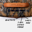 Close-up of the back of a TurtleBox speaker with a camouflage pattern, a tan handle, and black control buttons. Hand-drawn arrows point to the buttons with labels reading BLUETOOTH, 2-BOX STEREO PAIRING, and PARTY MODE.