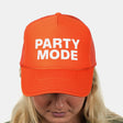 A woman wearing a bright orange trucker hat with the words 'PARTY MODE' printed in bold white letters on the front, against a light gray background.