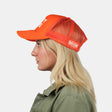 A woman in profile wearing a bright orange trucker hat with white lettering. The hat features a solid orange front panel and brim, with orange mesh back panels.