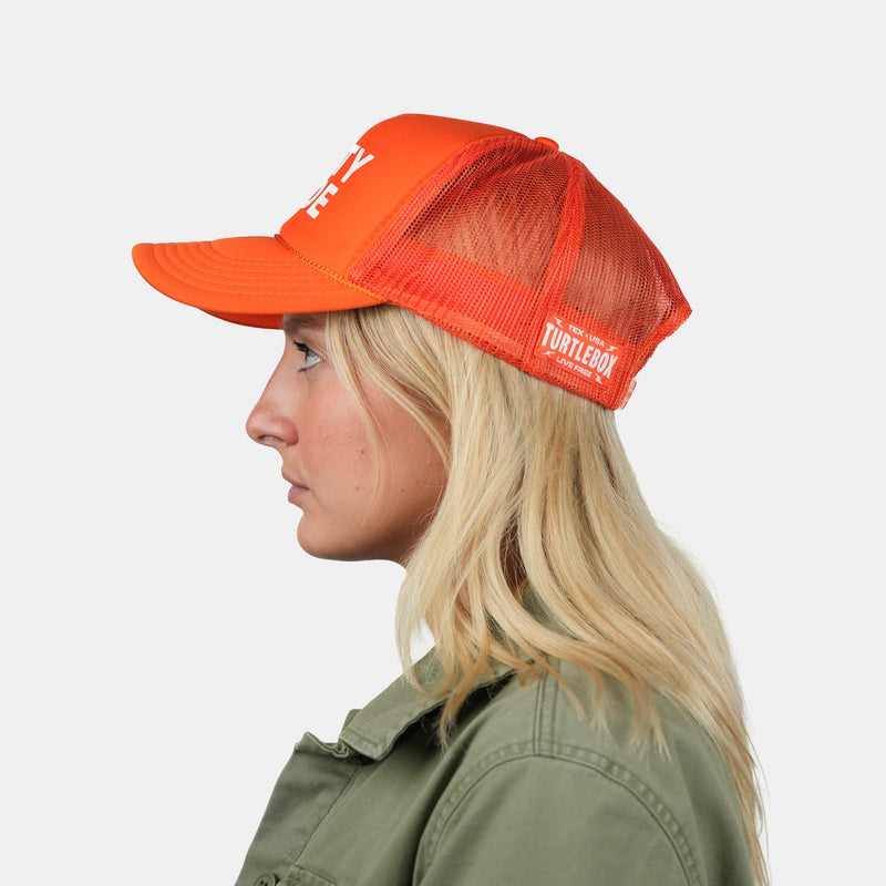 A woman in profile wearing a bright orange trucker hat with white lettering. The hat features a solid orange front panel and brim, with orange mesh back panels.