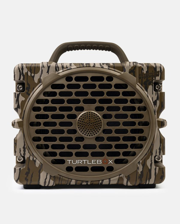 A rugged portable speaker with a camouflage pattern in earthy brown, beige, and green tones. It features a large circular front grille, a top carrying handle, and the 'TURTLEBX' logo printed on the front.