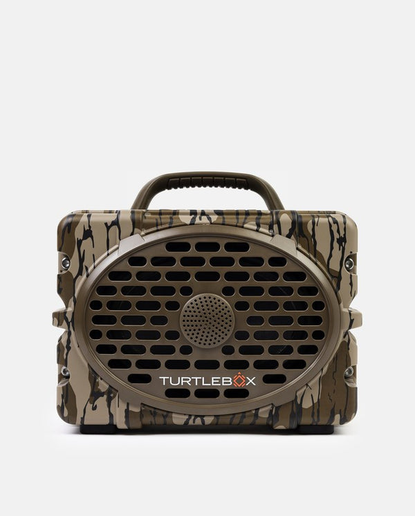 A portable speaker by TurtleBox featuring a camouflage pattern in shades of brown, beige, and green, with a dark brown oval speaker grille and handle on a light gray background.