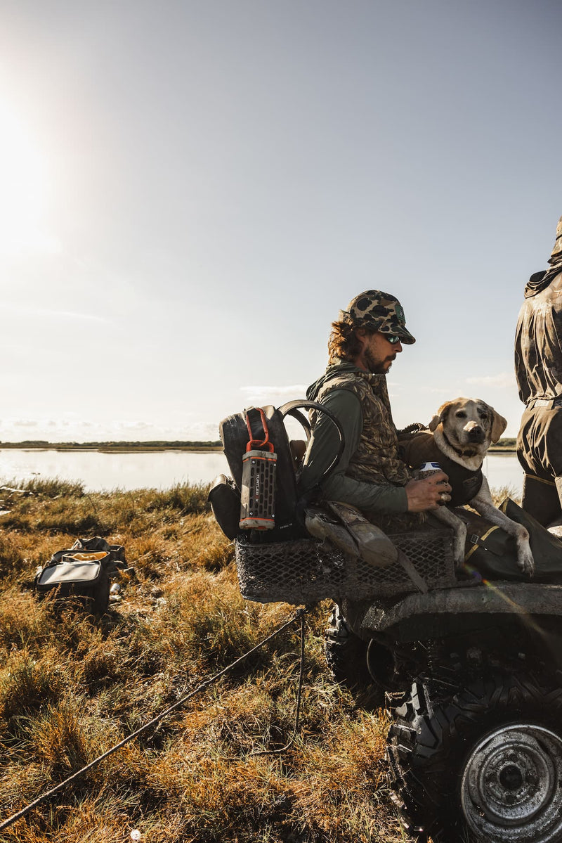 A lifestyle photo of two hunters with a dog on an ATV near a body of water with a Delta Turtlebox Ranger speaker attached to a backpack. #base-color_delta