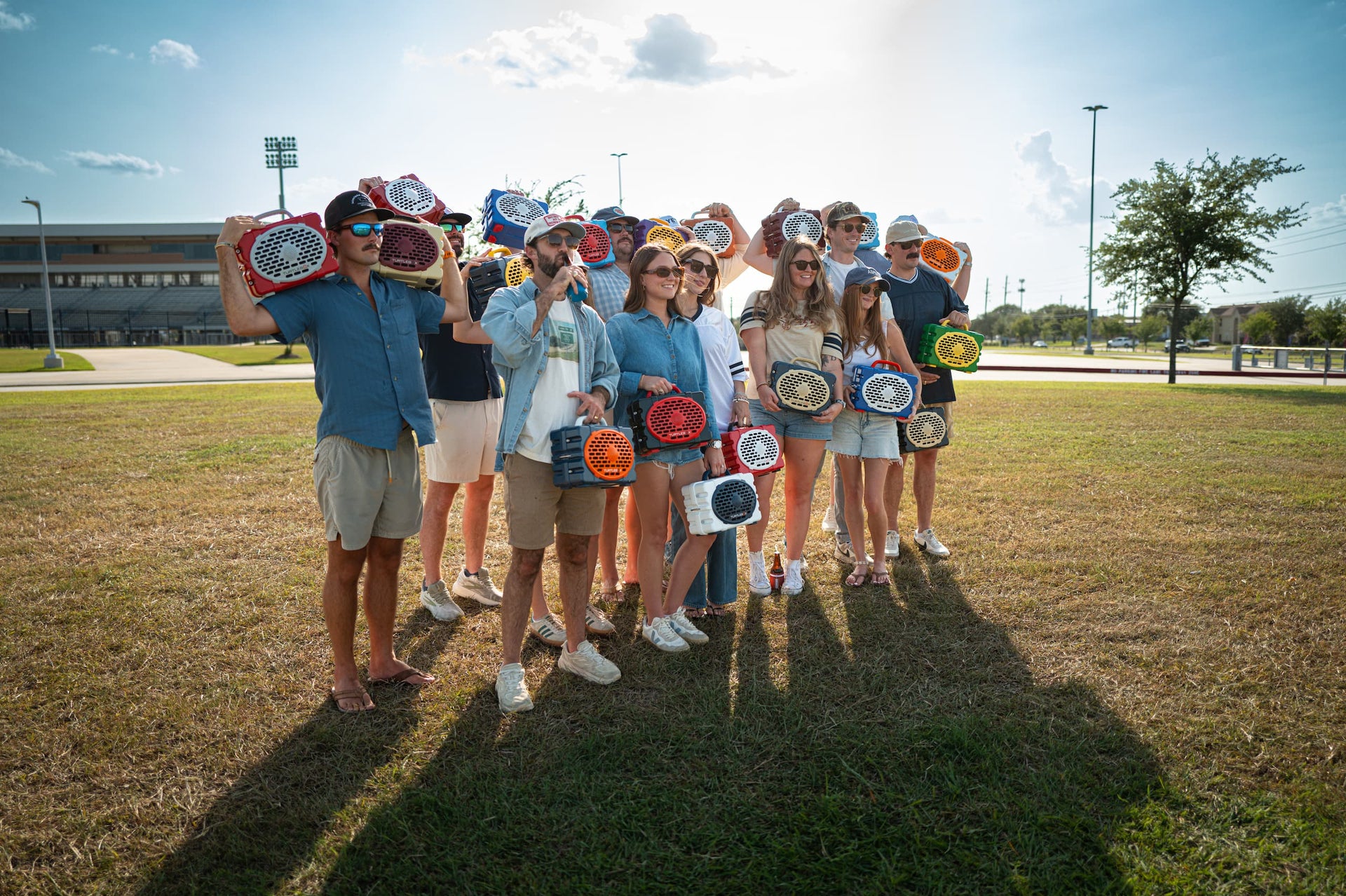 A group of young people standing outdoors on a grassy field, each holding a small round award plaque. The group is photographed in bright daylight with trees, stadium lights, and a building visible in the background.