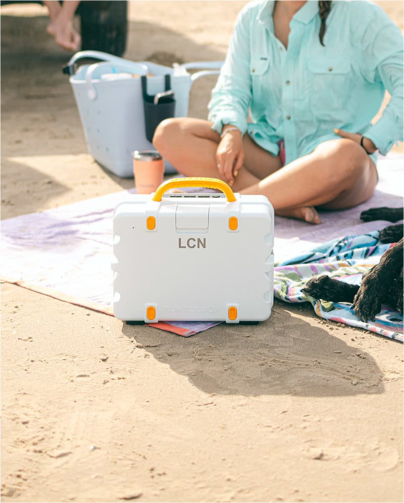 A woman in a light blue shirt and blue cap sits on a blanket on a sandy beach, with a white cooler in front of her and a black dog lying nearby. A man is seated on the back of a truck in the background, along with another cooler and an orange tote bag.