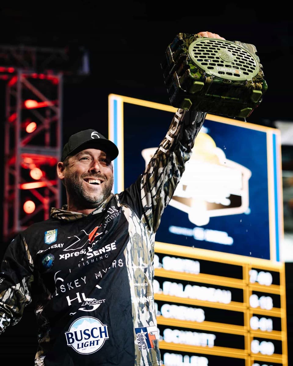 A smiling man in a black and white patterned long-sleeve shirt and a black cap raises a large green camouflage speaker above his head in a celebratory moment at night, with a scoreboard and stage lights in the background.