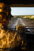A person in camouflage clothing sits inside an off-road vehicle, looking out a windshield toward a dirt path through a grassy landscape. A yellow and brown camouflage speaker sits on the vehicle's rack.