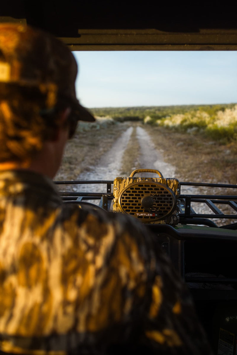 A person in camouflage clothing sits inside an off-road vehicle, looking out a windshield toward a dirt path through a grassy landscape. A yellow and brown camouflage speaker sits on the vehicle's rack.