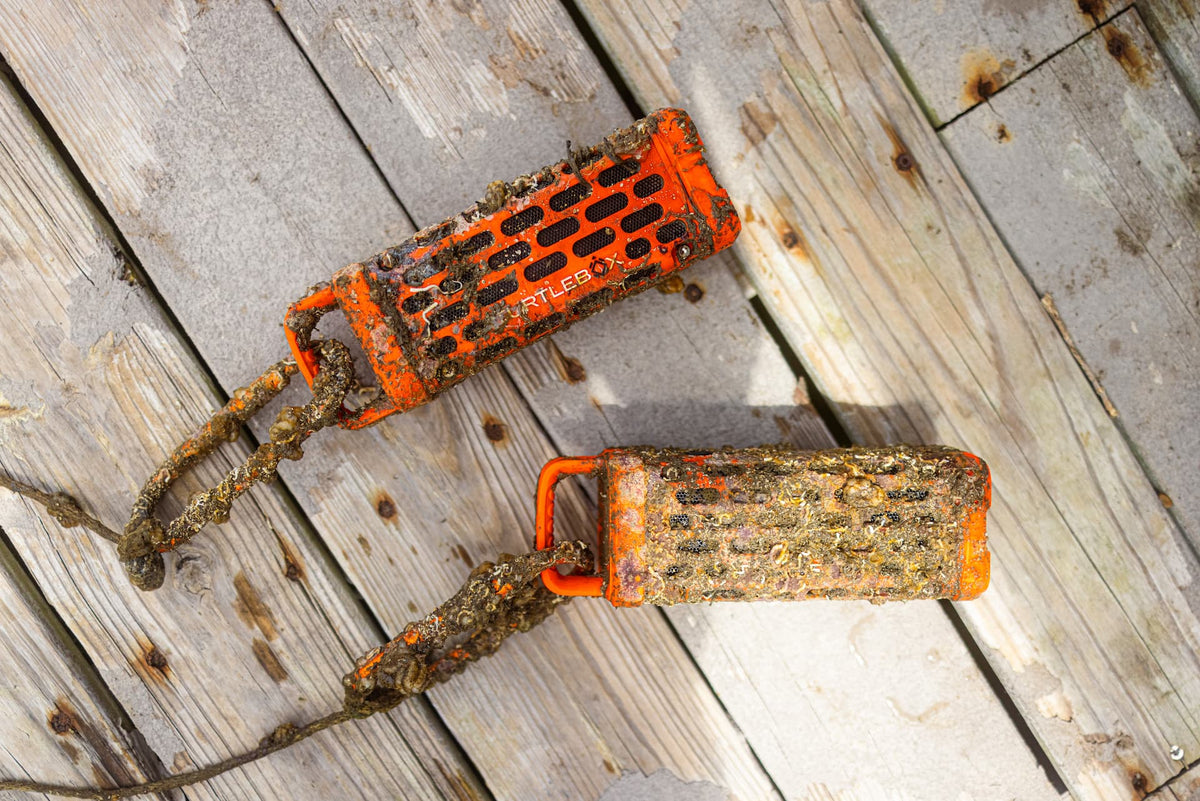 Two orange Ranger speakers covered in mud and barnacles, resting on weathered wooden planks.