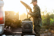 A lifestyle photo of a person in camouflage holding a bow, with a TURTLE BECK power station on a truck bed in a natural setting. #base-color_gray