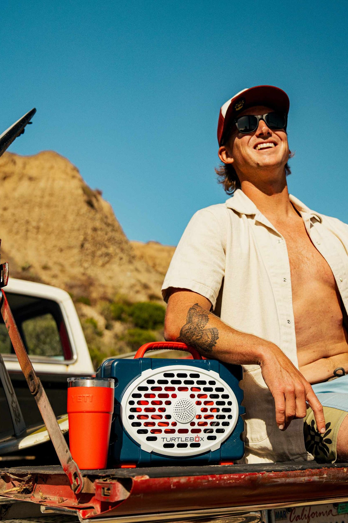 A lifestyle photo of a man sitting in the back of a pickup truck with a vintage radio, mountains in the background #speaker-color_americana-blue