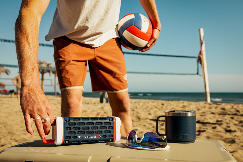 A lifestyle photo of a person on a beach with a volleyball, cooler, and sunglasses, featuring a TURTLE BECK product #base-color_americana-blue