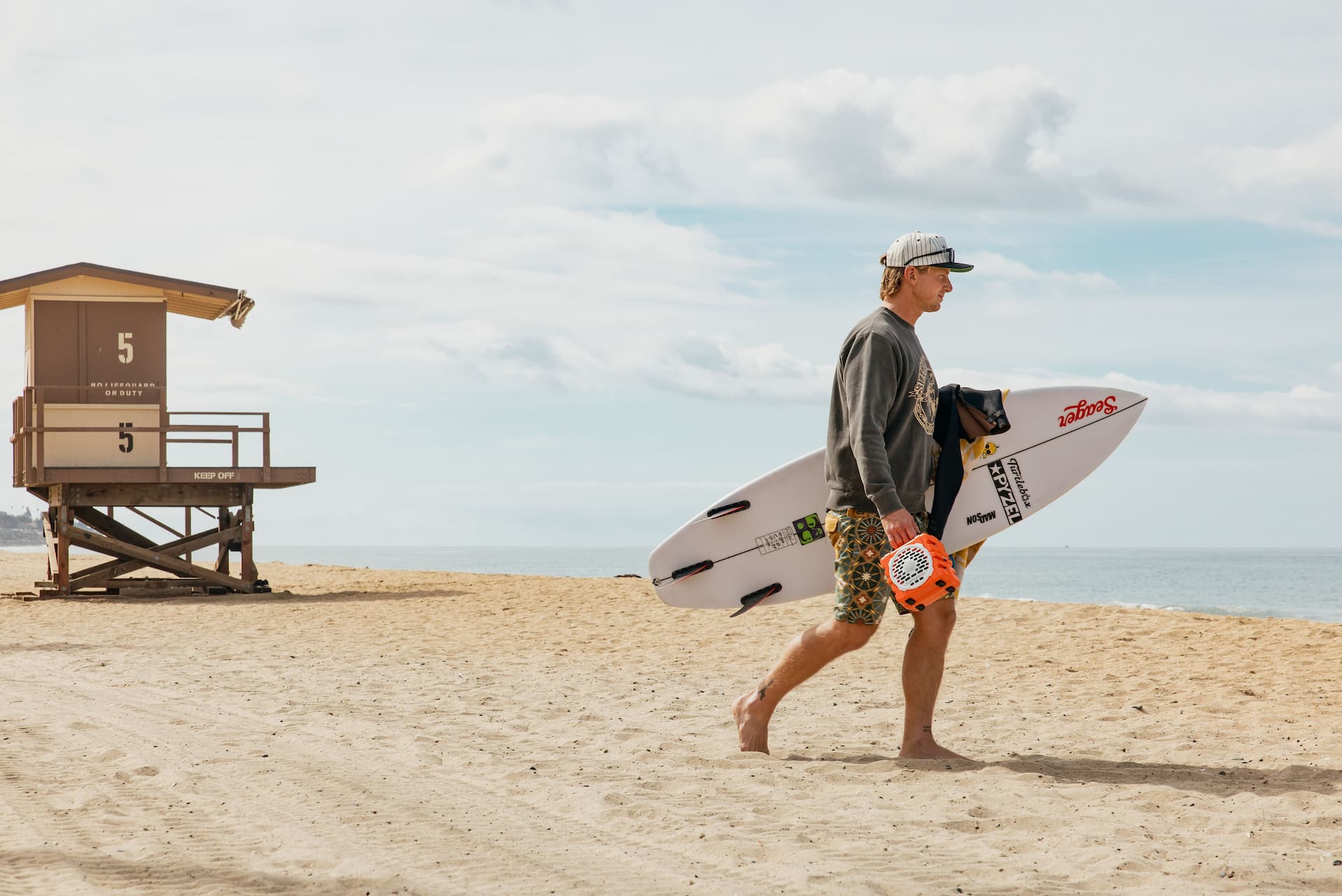 Man walking on a beach with a surfboard, holding an Turtlebox Cub Orange speaker.