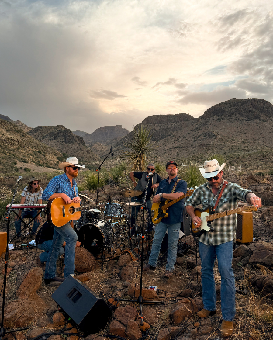 A group of five musicians performing live outdoor acoustic music on a rocky desert mountain landscape at sunset. The band members, including guitarists, a keyboardist, and a drummer, are dressed in casual western and blue-collar attire with cowboy hats.