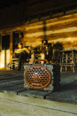 A rugged portable speaker with a camouflage pattern and copper-colored front grille sits on a wooden porch in front of a log cabin, with two people blurred in the background.