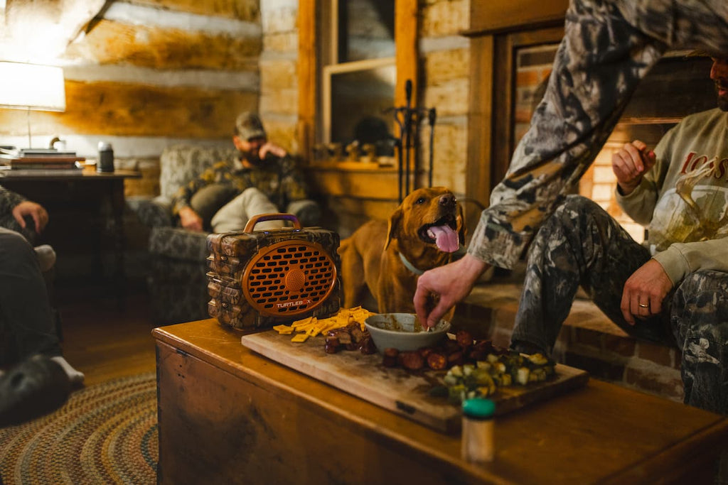 A warm, rustic indoor scene with people and a dog gathered around a wooden table. In the foreground, a hand reaches toward a bowl on a cutting board with food, while another person in camouflage sits nearby.
