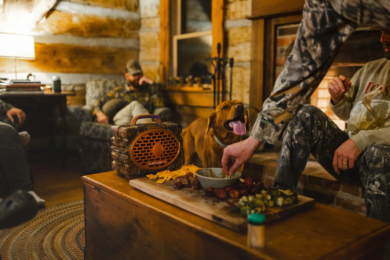 A warm, rustic indoor scene with people and a dog gathered around a wooden table. In the foreground, a hand reaches toward a bowl on a cutting board with food, while another person in camouflage sits nearby.