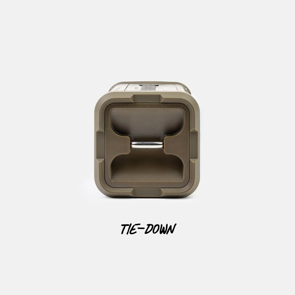 A top-down view of a square outdoor storage container lid in a muted olive-tan color, featuring a central tie-down loop and the text 'TIE-DOWN' printed below on a light gray background.