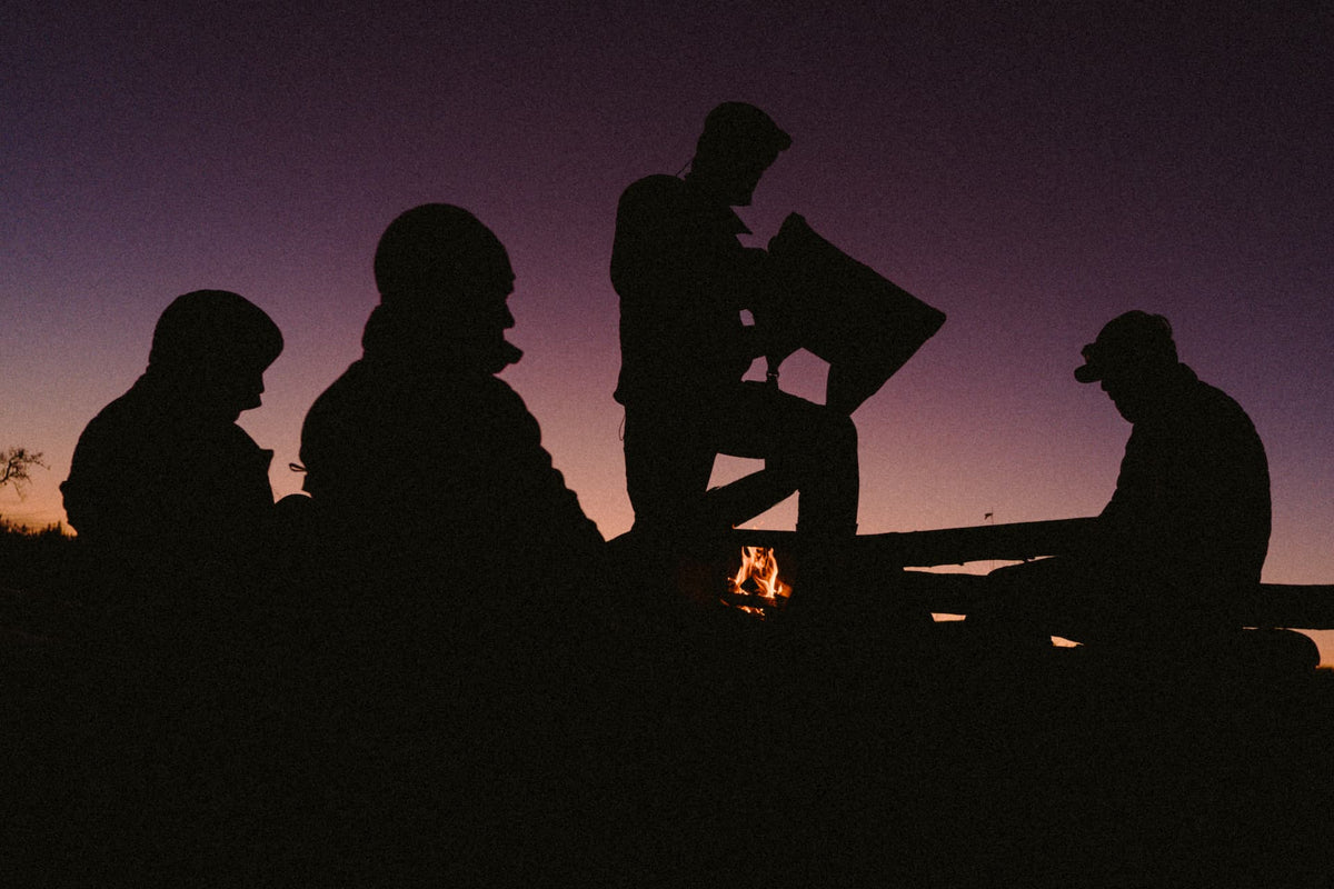 Silhouetted figures gathered around a small fire at dusk against a purple and pink sky, with one person holding a rectangular object above the flames.