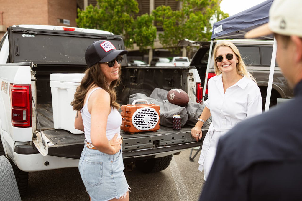 A lifestyle photo of two women standing next to a pickup truck with sports equipment in the bed, in an outdoor setting. #speaker-color_burnt-orange-white-gameday-series