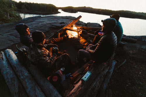 People sitting around a campfire by a body of water at sunset.
