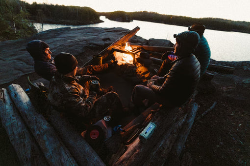 People sitting around a campfire by a body of water at sunset.