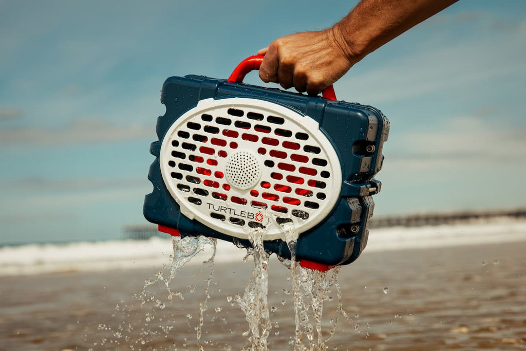 A lifestyle photo of a person holding a waterproof device labeled 'Turtle Beach' on a beach with water splashing out. #speaker-color_americana-blue