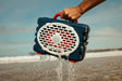 A lifestyle photo of a person holding a waterproof device labeled 'Turtle Beach' on a beach with water splashing out. #speaker-color_americana-blue