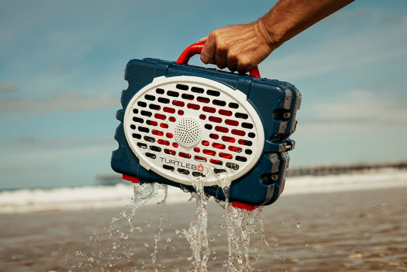 A lifestyle photo of a person holding a waterproof device labeled 'Turtle Beach' on a beach with water splashing out. #speaker-color_americana-blue