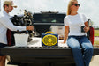 A lifestyle photo of a Original Gen 3 Navy and yellow portable speaker sits in the bed of a pickup truck, with a man loading portable chairs on the left and a woman sitting on the tailgate on the right.