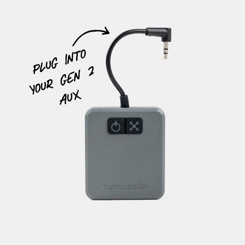 A TurtleBox audio adapter device in gray with a black 3.5mm jack cable, shown on a light gray background with a handwritten note indicating how to plug it into a Gen 2 AUX port.