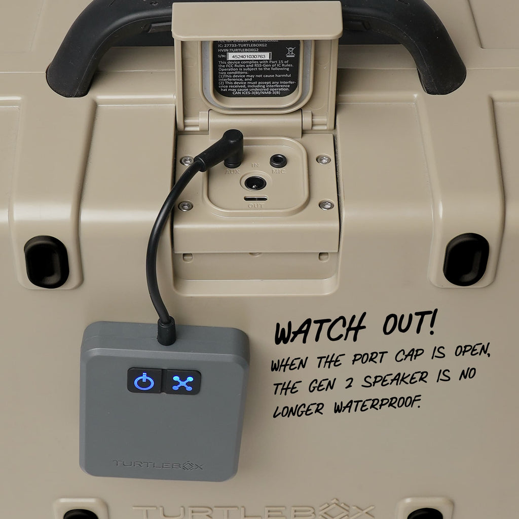 Close-up of a TURTLEBOX cooler showing the rear port area with a gray power adapter plugged into the Gen 2 speaker port. A black warning label is visible inside the port cap, and handwritten text on the right reads 'WATCH OUT! WHEN THE PORT CAP IS OPEN, THE GEN 2 SPEAKER IS NO LONGER WATERPROOF.