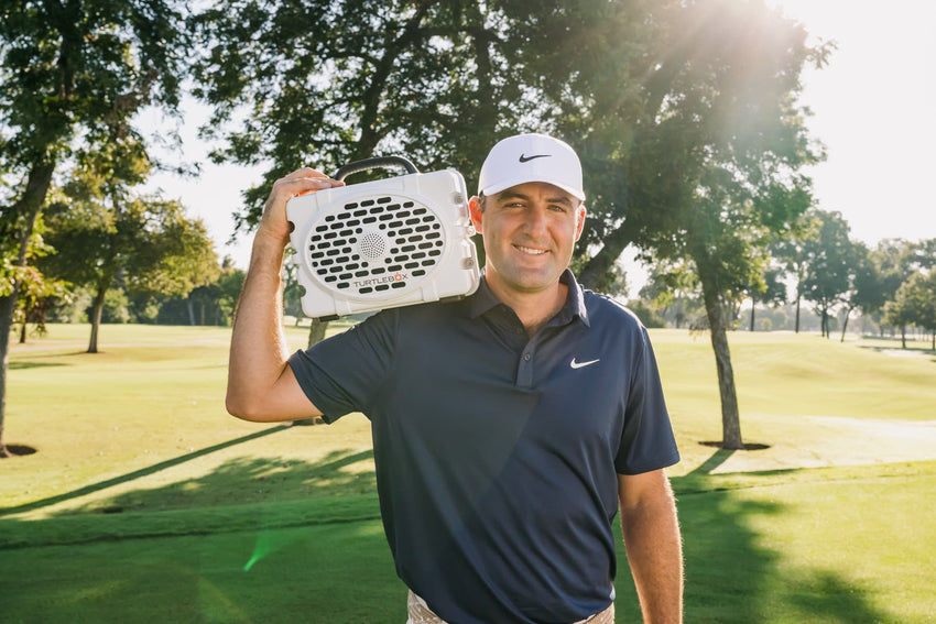 Scottie Scheffler holding a white Original Turtlebox speaker on a golf course with trees in the background