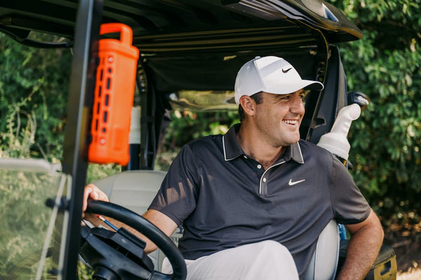 Scottie Scheffler driving a golf cart with a golf club, wearing a Nike cap and shirt.