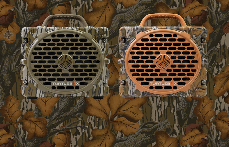 Two TurtleBox portable speakers shown side by side against a woodland camouflage background. The left speaker has a beige and brown bark camo pattern, and the right speaker has an orange and brown bark camo pattern.