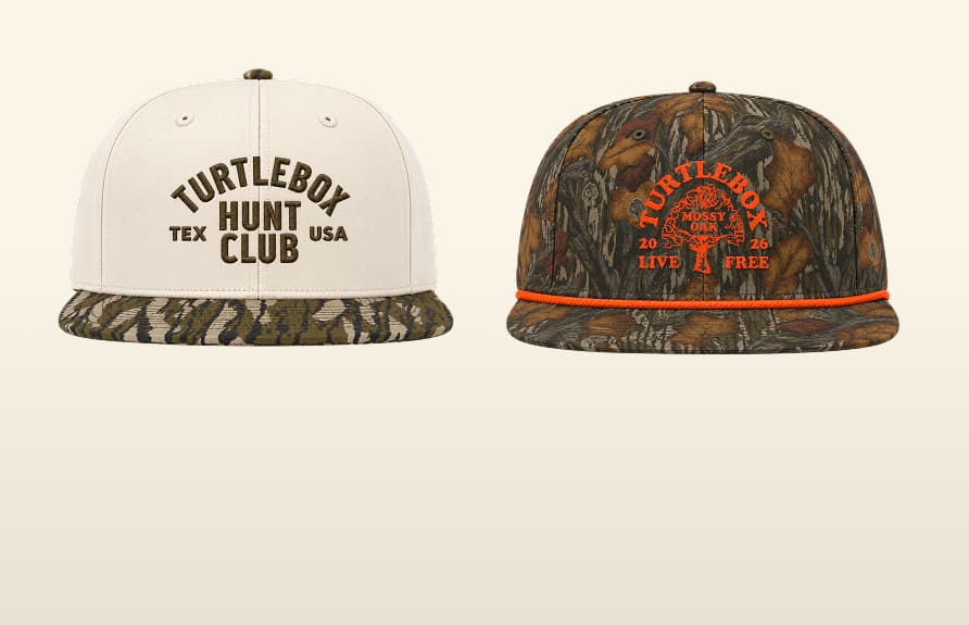 Two Turtlebox baseball caps displayed against a camouflage leaf and tree bark background. The left cap is off-white with brown embroidered text reading 'TURTLEBOX TEXAS HUNT CLUB USA' and a matching camouflage brim.