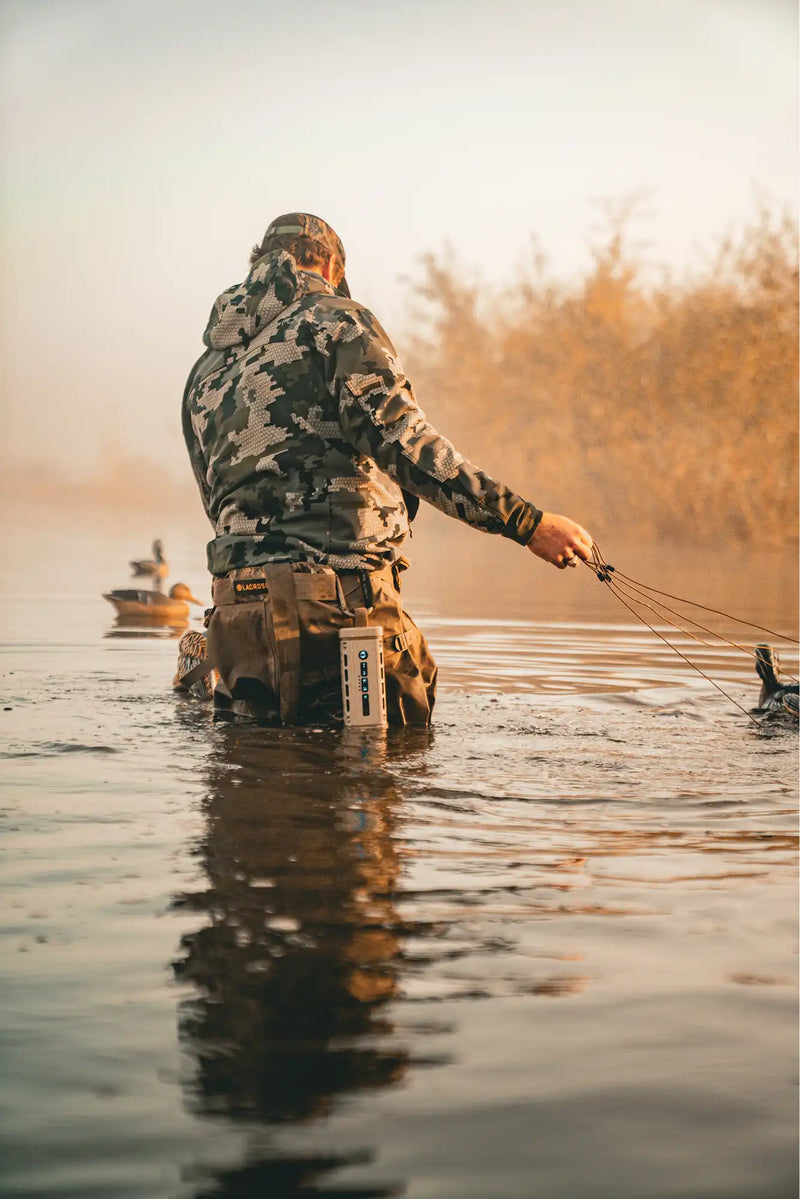Person in camouflage wading through water with a fishing rod, sunset or sunrise in the background.