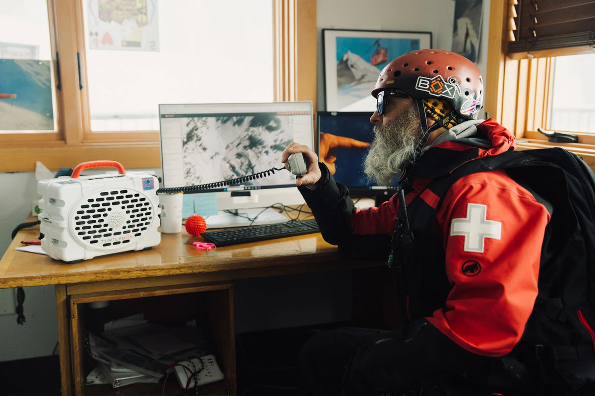 Person in a red jacket with a white cross sitting at a desk using a computer, wearing a helmet and goggles.