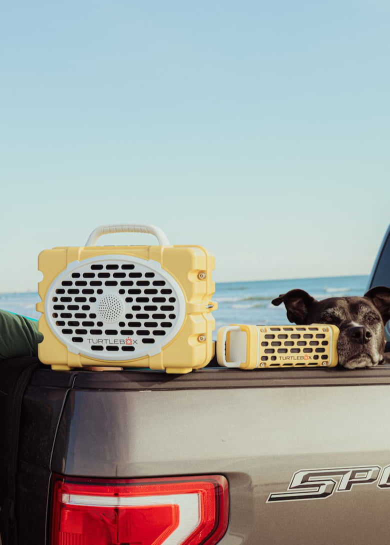 Yellow portable speaker on a car's open trunk with a clear blue sky and ocean in the background.