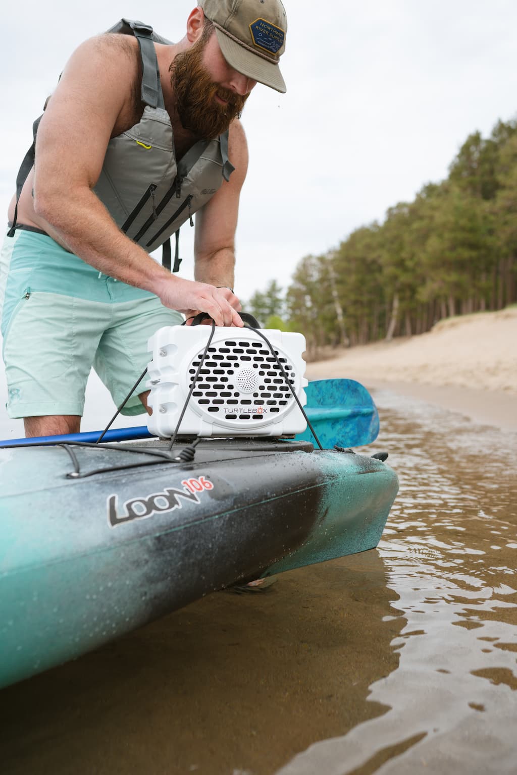 A lifestyle photo of a bearded guy with a vest and a hat putting a white speaker in a kayak #speaker-color_white