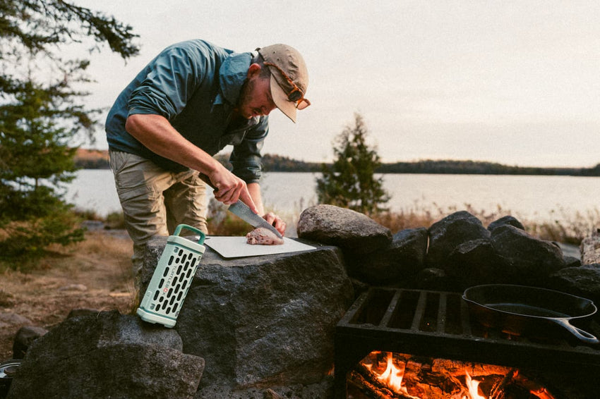 A lifestyle photo showing a man cutting meat next to an open camp fire with the River Rock Ranger speaker leaning against a rock. #base-color_river-rock